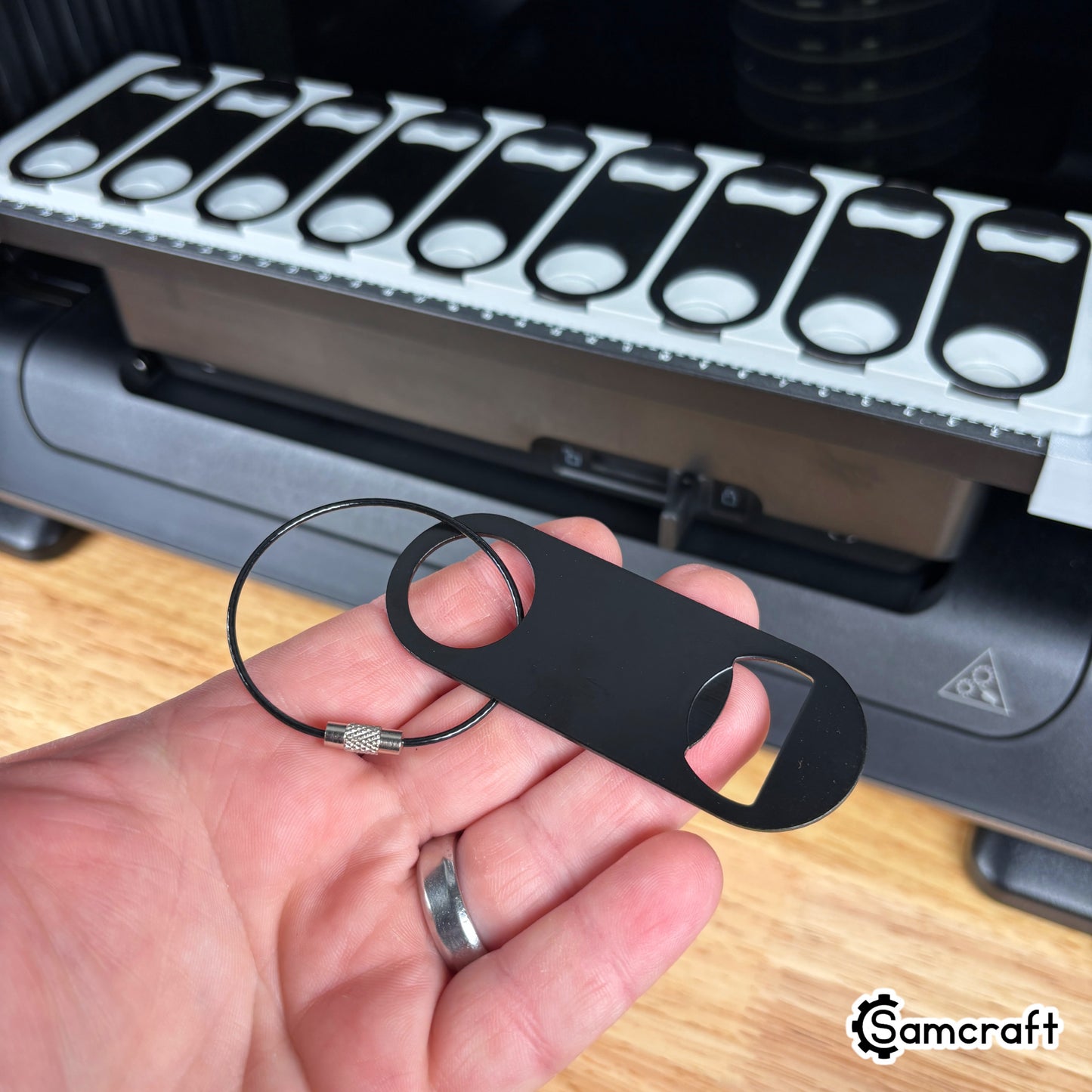 Oval Metal Bottle Opener Keychain Jig for the eufyMake E1 — hand holding a single black oval metal bottle opener with keychain ring beside the loaded gray 3D-printed tray, showing the blank to be printed.