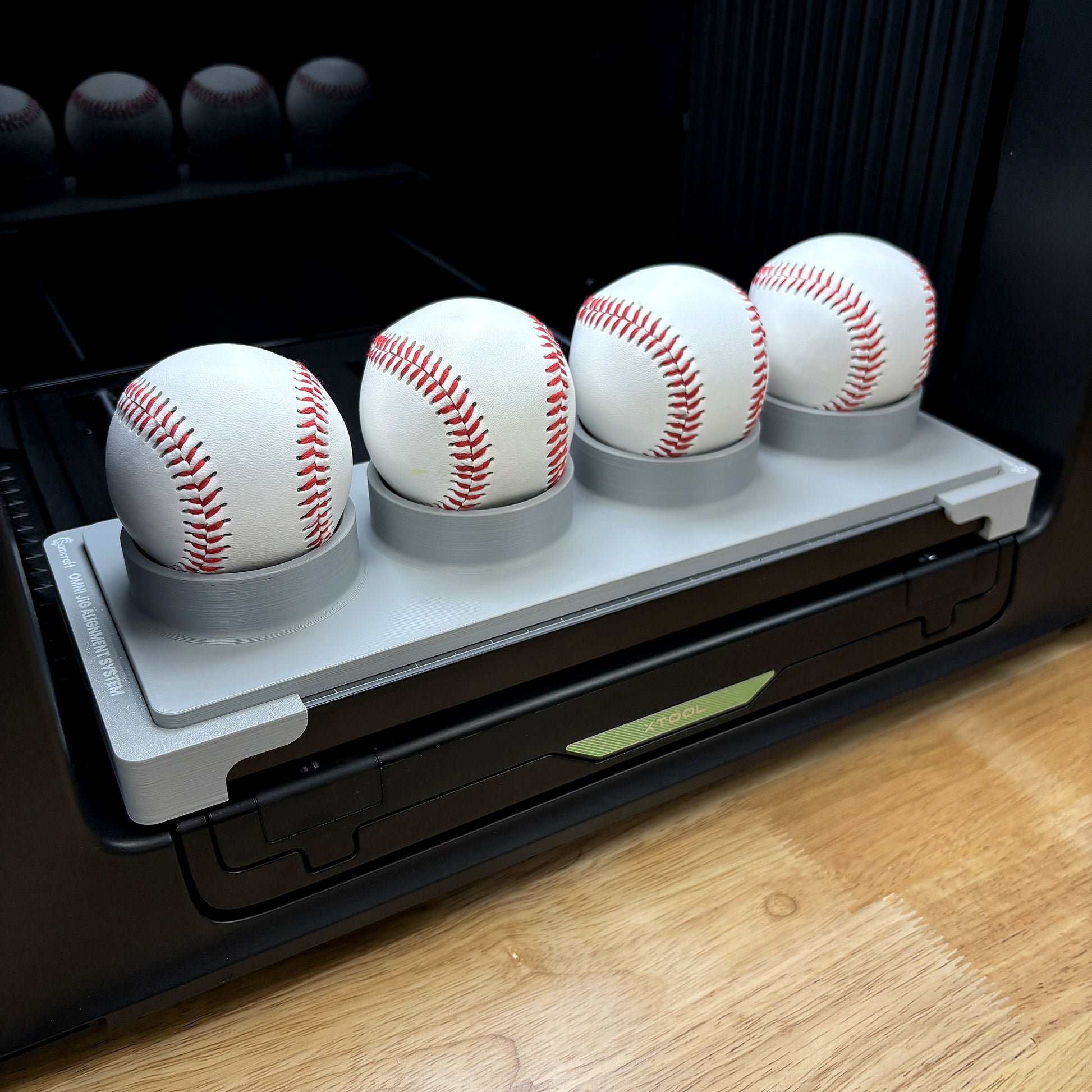 Samcraft baseball, softball and pickleball jig on the bed of an xTool Omni UV printer, holding four white baseballs in raised spherical cradles.