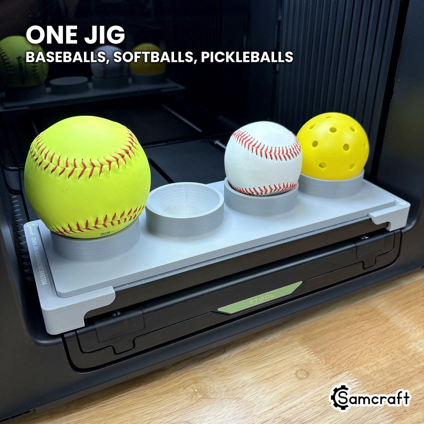 A yellow softball, a white baseball and a yellow pickleball all seated in the same Samcraft jig on the xTool Omni bed, showing one jig covers three ball types.