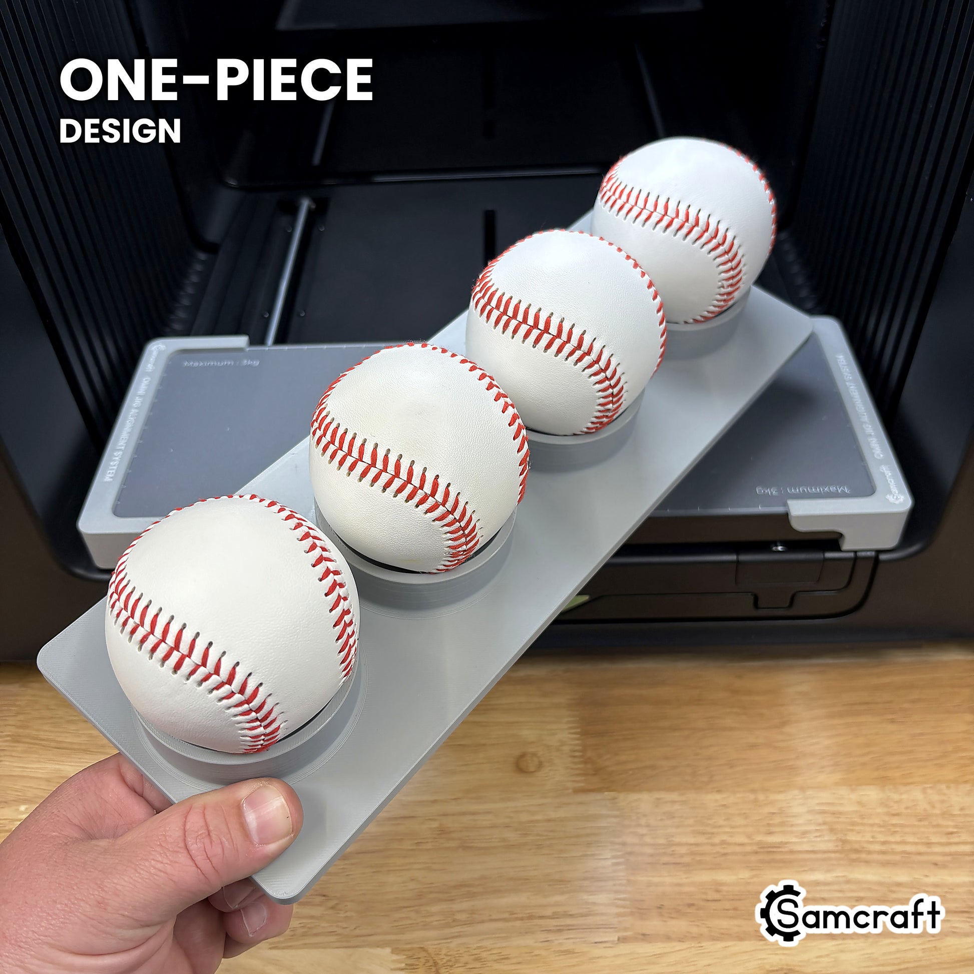 Hand holding the one-piece Samcraft baseball jig for the xTool Omni, four baseballs seated in a single molded gray tray.