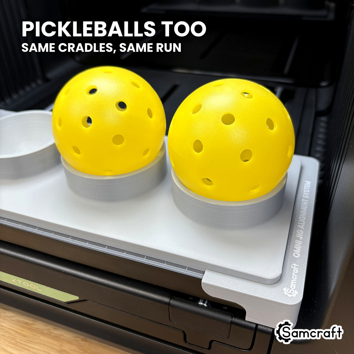 Two yellow pickleballs seated in the Samcraft jig on the xTool Omni printer bed, using the same cradles as the baseballs.