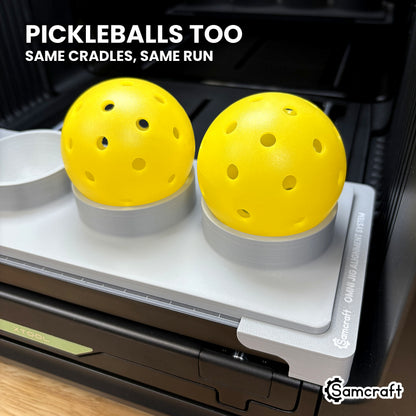 Two yellow pickleballs seated in the Samcraft jig on the xTool Omni printer bed, using the same cradles as the baseballs.