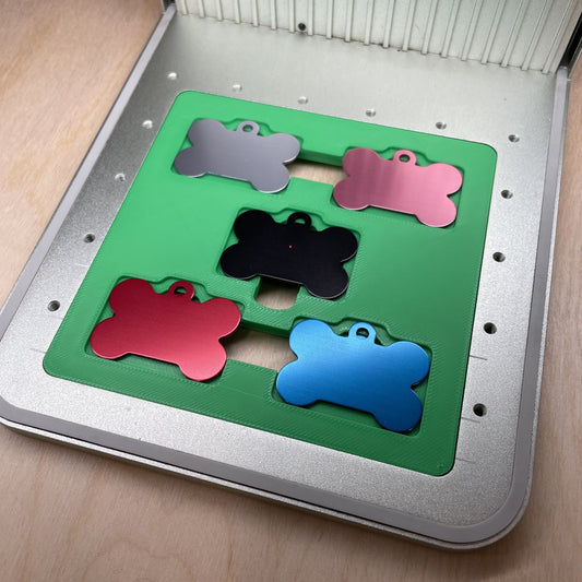 Samcraft bone pet tag jig in an xTool F1 laser bed, holding five bone-shaped aluminium pet tags in grey, pink, black, red and blue.