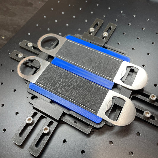 Samcraft blue large bottle opener jig on a ComMarker laser bed, holding a black leather-wrapped bottle opener and two steel openers.