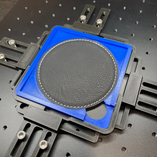 Samcraft blue drink coaster jig on a ComMarker laser bed, holding a round dark leatherette coaster with a stitched edge.