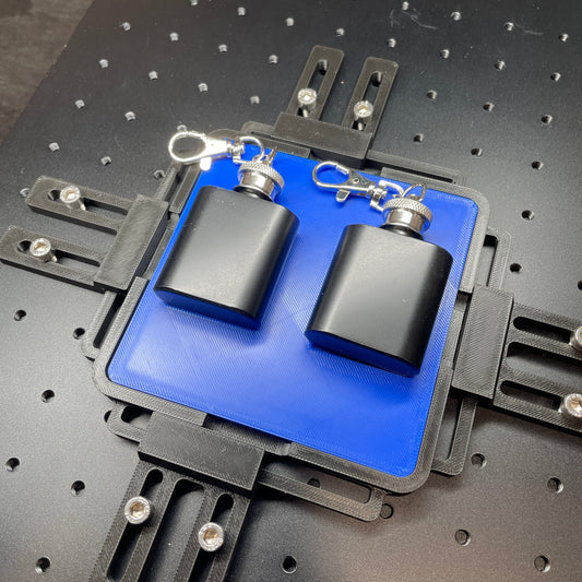 Samcraft blue flask keychain jig on a ComMarker laser bed, holding two black flask keychains.