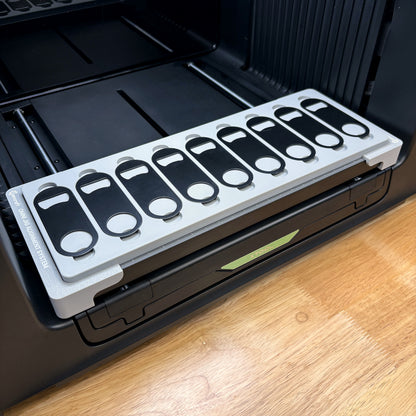 Samcraft bottle opener jig on the bed of an xTool Omni UV printer, holding nine flat metal keychain openers in a single row.