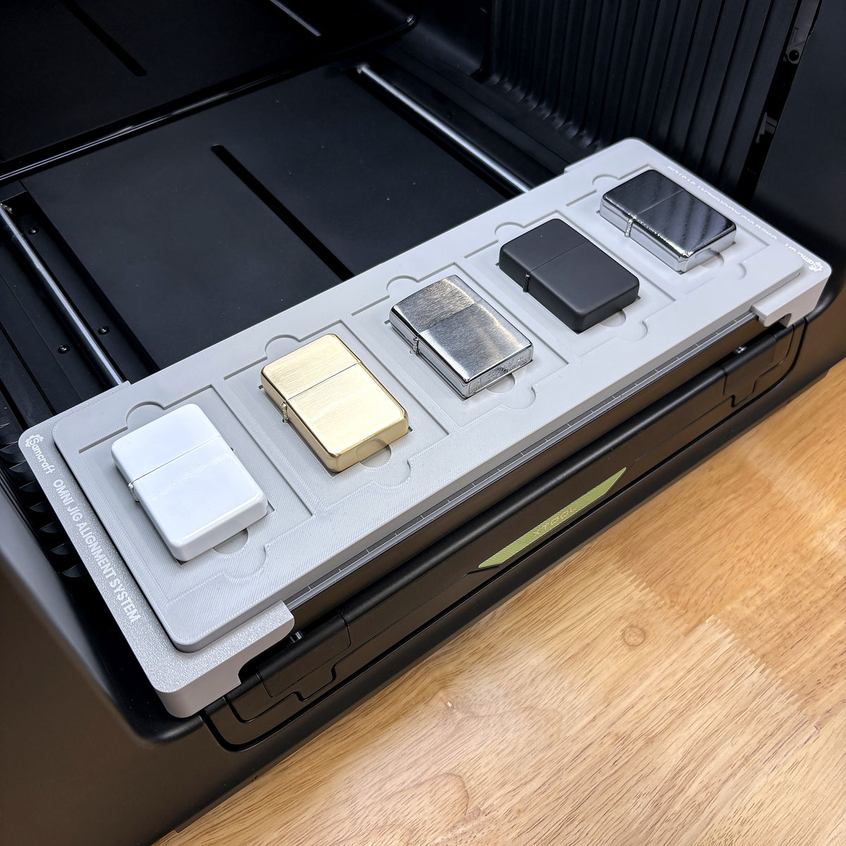 Samcraft flip top lighter jig on the bed of an xTool Omni UV printer, holding five lighters in brass, chrome, matte black and satin finishes.