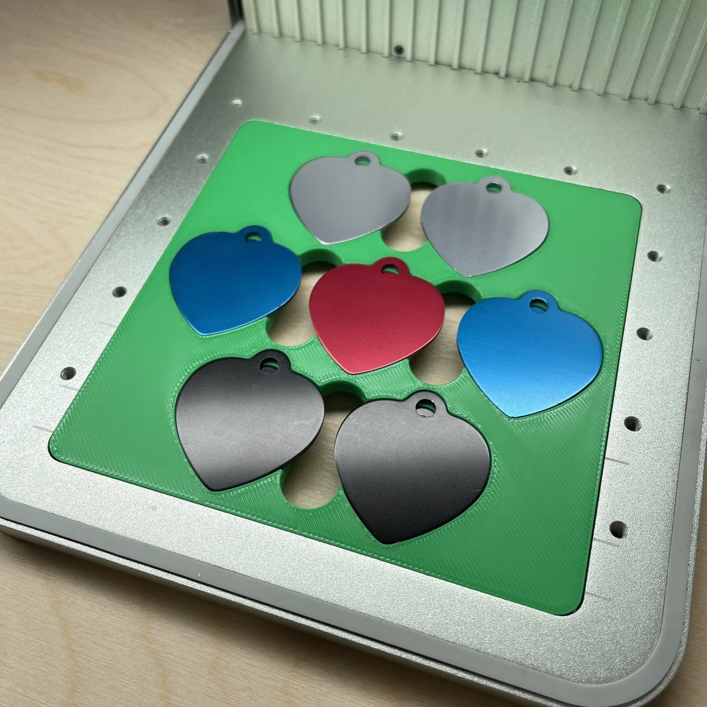 Samcraft heart pet tag jig in an xTool F1 laser bed, holding seven heart-shaped aluminium pet tags in grey, blue, red and black.