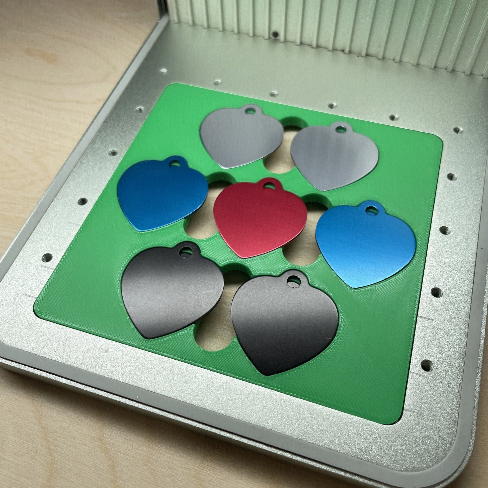 Samcraft heart pet tag jig in an xTool F1 laser bed, holding seven heart-shaped aluminium pet tags in grey, blue, red and black.