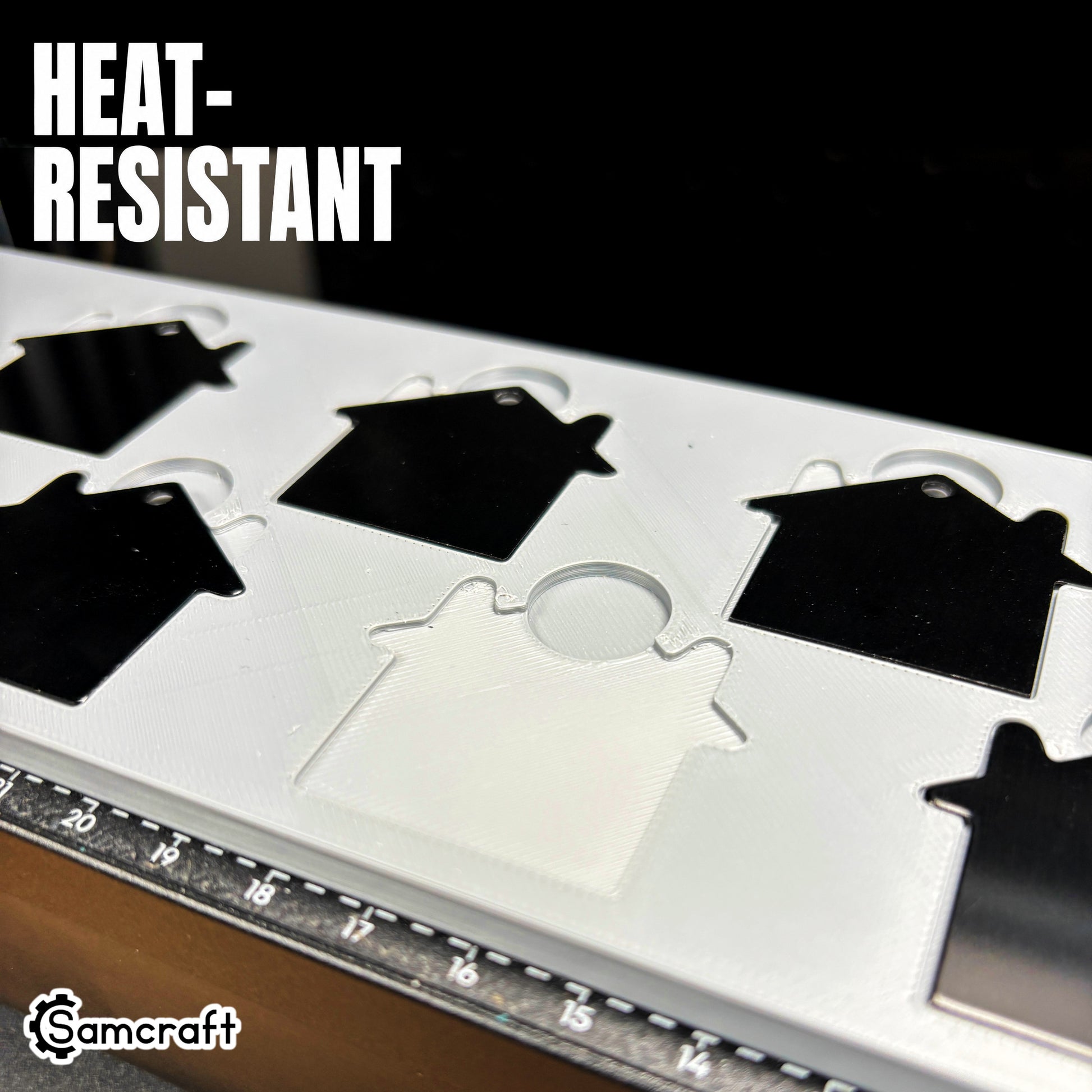 Close-up of black house-shaped keychain blanks seated in the Samcraft jig's heat-resistant gray tray with house-shaped recessed pockets and alignment ruler on the eufyMake E1.