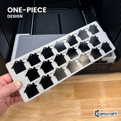 Hand holding the one-piece Samcraft house tag keychain jig for the xTool Omni, twenty-one tags seated in a single molded tray.