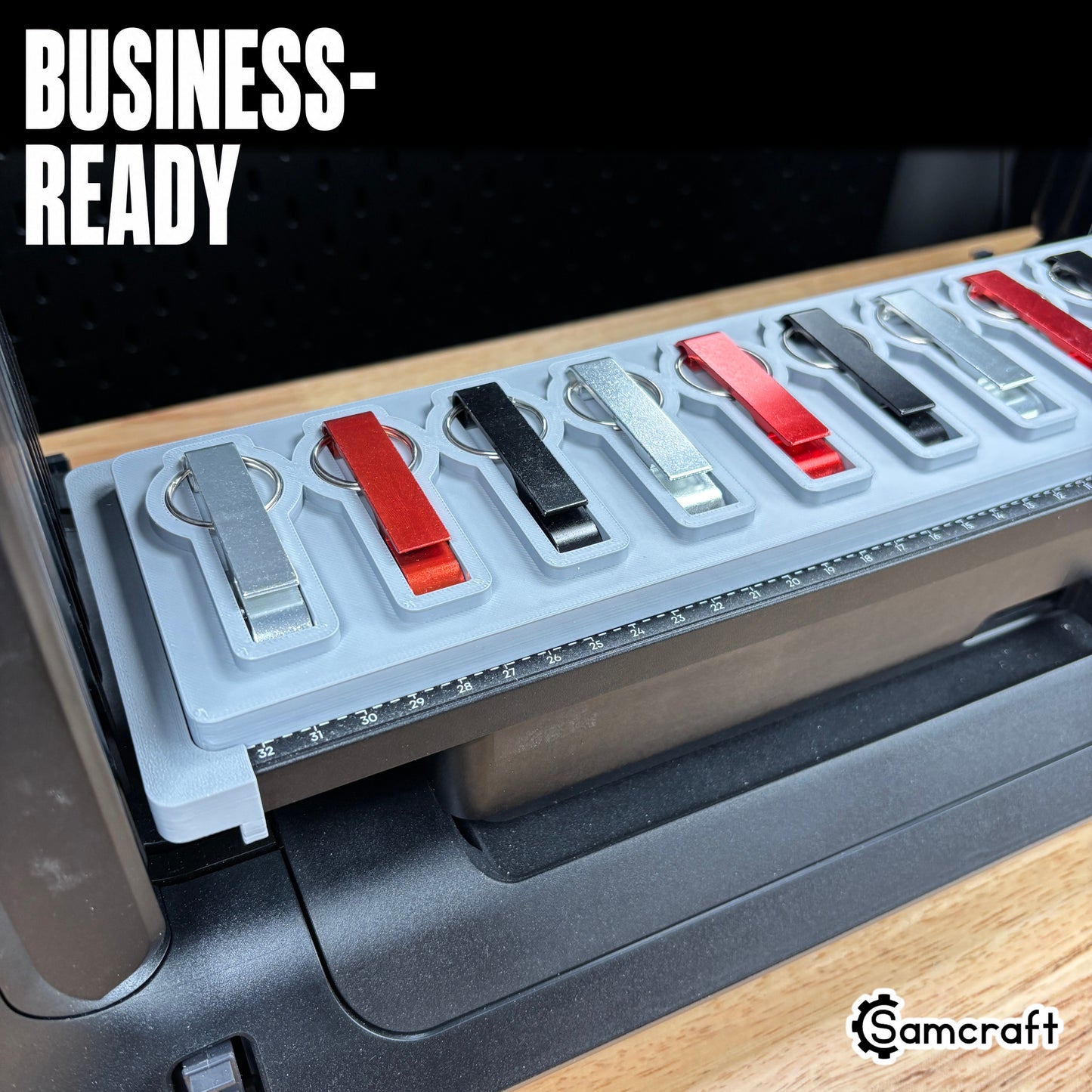 Keychain Bottle Opener Jig for the eufyMake E1 — angled view of silver, red, and black metal keychain bottle openers with split rings seated in the gray 3D-printed tray with built-in alignment ruler, ready for business-scale personalization.