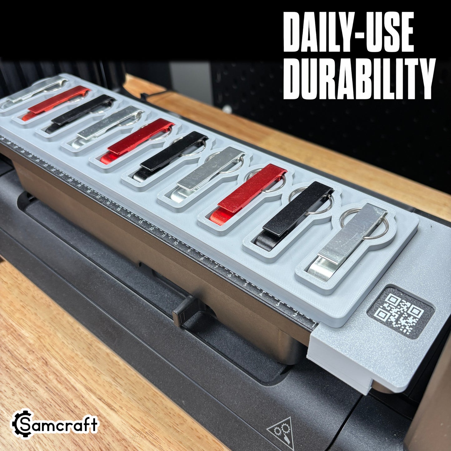 Keychain Bottle Opener Jig for the eufyMake E1 — metal keychain bottle openers in silver, red, and black finishes resting in the shaped pockets of the gray one-piece tray with built-in alignment ruler and QR code, built for daily-use durability.