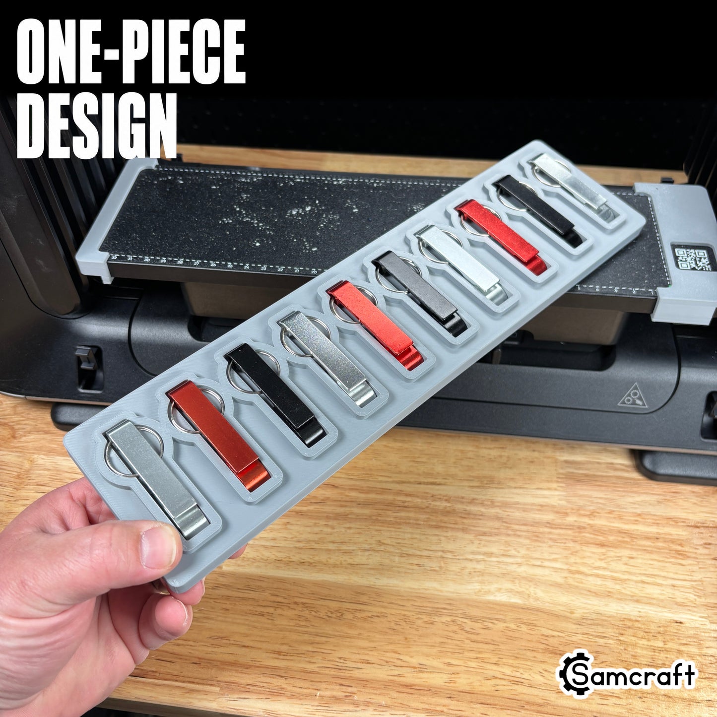 Keychain Bottle Opener Jig for the eufyMake E1 — hand holding the gray one-piece 3D-printed tray loaded with silver, red, and black metal keychain bottle openers in their molded pockets, showing the strong single-piece design.