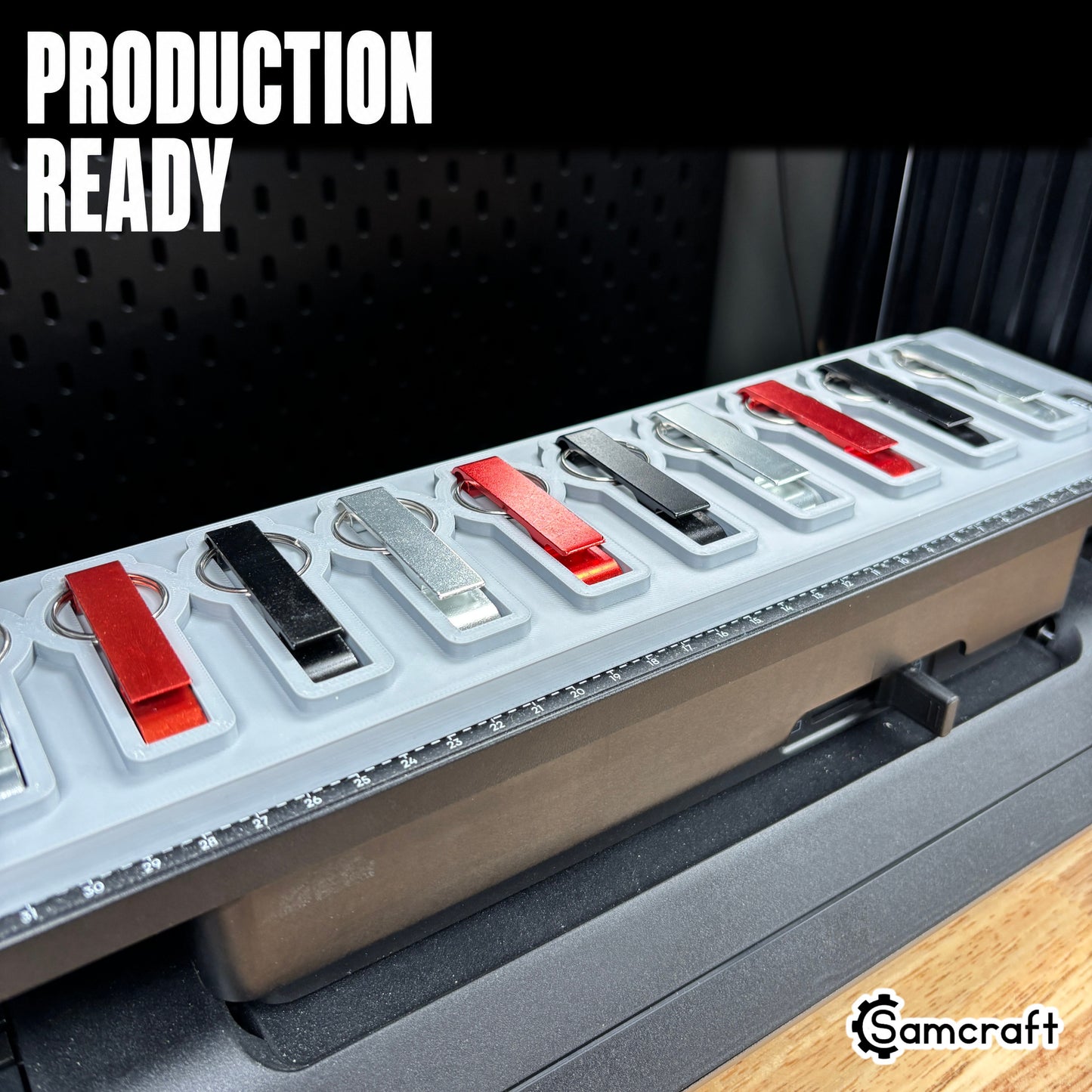Keychain Bottle Opener Jig for the eufyMake E1 — a row of red, black, and silver metal keychain bottle openers with split rings lined up in the gray one-piece tray mounted on the printer bed, alignment ruler along the edge, set up for production-ready batch printing.