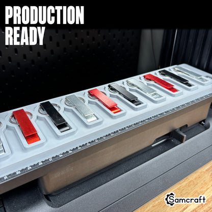 Keychain Bottle Opener Jig for the eufyMake E1 — a row of red, black, and silver metal keychain bottle openers with split rings lined up in the gray one-piece tray mounted on the printer bed, alignment ruler along the edge, set up for production-ready batch printing.