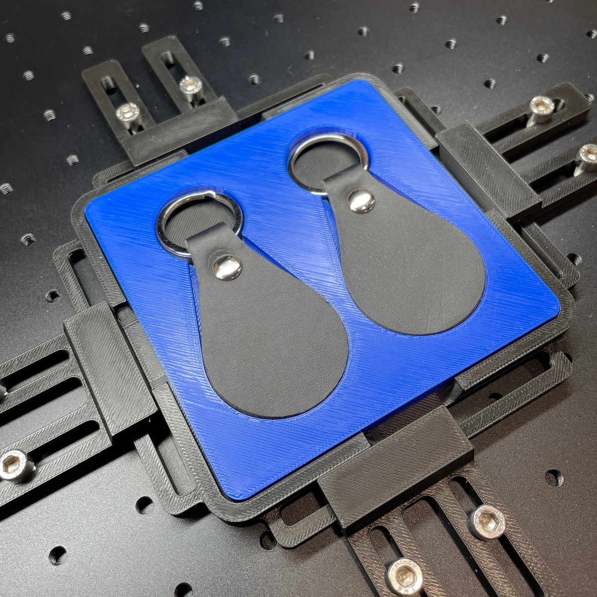 Samcraft blue leather key fob jig on a ComMarker laser bed, holding two black teardrop leather key fobs.