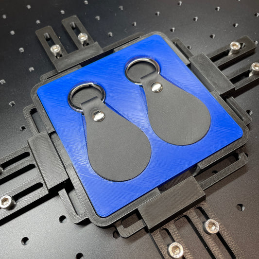 Samcraft blue leather key fob jig on a ComMarker laser bed, holding two black teardrop leather key fobs.