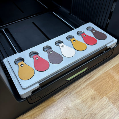 Samcraft leather key fob jig on the bed of an xTool Omni UV printer, holding seven teardrop leather fobs in tan, red, brown and cream.
