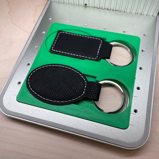 Samcraft green leatherette keychain jig in an xTool F1 laser bed, holding a black rectangular and a black oval leatherette keychain on split rings.