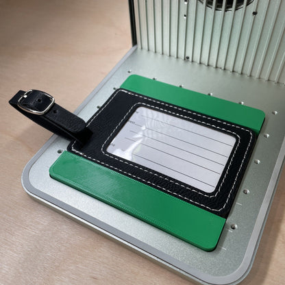 Samcraft leatherette luggage tag jig in an xTool F1 laser bed, holding a black leatherette luggage tag with its clear address window facing up.