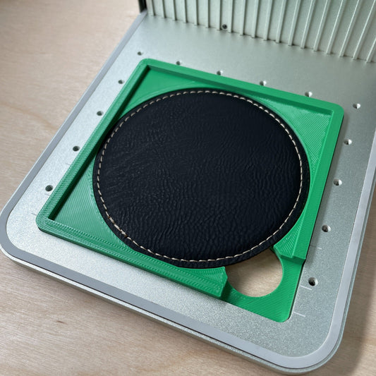 Samcraft drink coaster jig in an xTool F1 laser bed, holding a round black leatherette coaster.