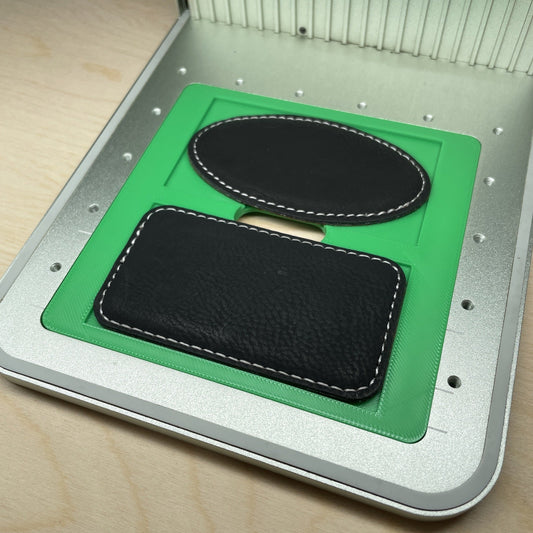 Samcraft magnetic badge jig in an xTool F1 laser bed, holding a black leatherette oval badge above a rectangular badge.
