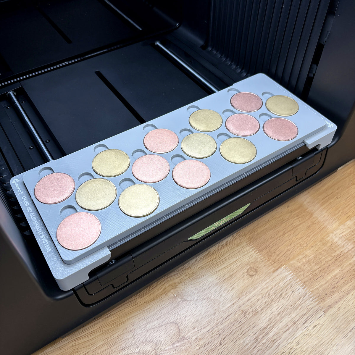 Samcraft 40mm metal coin jig on the bed of an xTool Omni UV printer, holding fifteen rose gold and gold blank challenge coins, seated on the Omni Jig Alignment System.