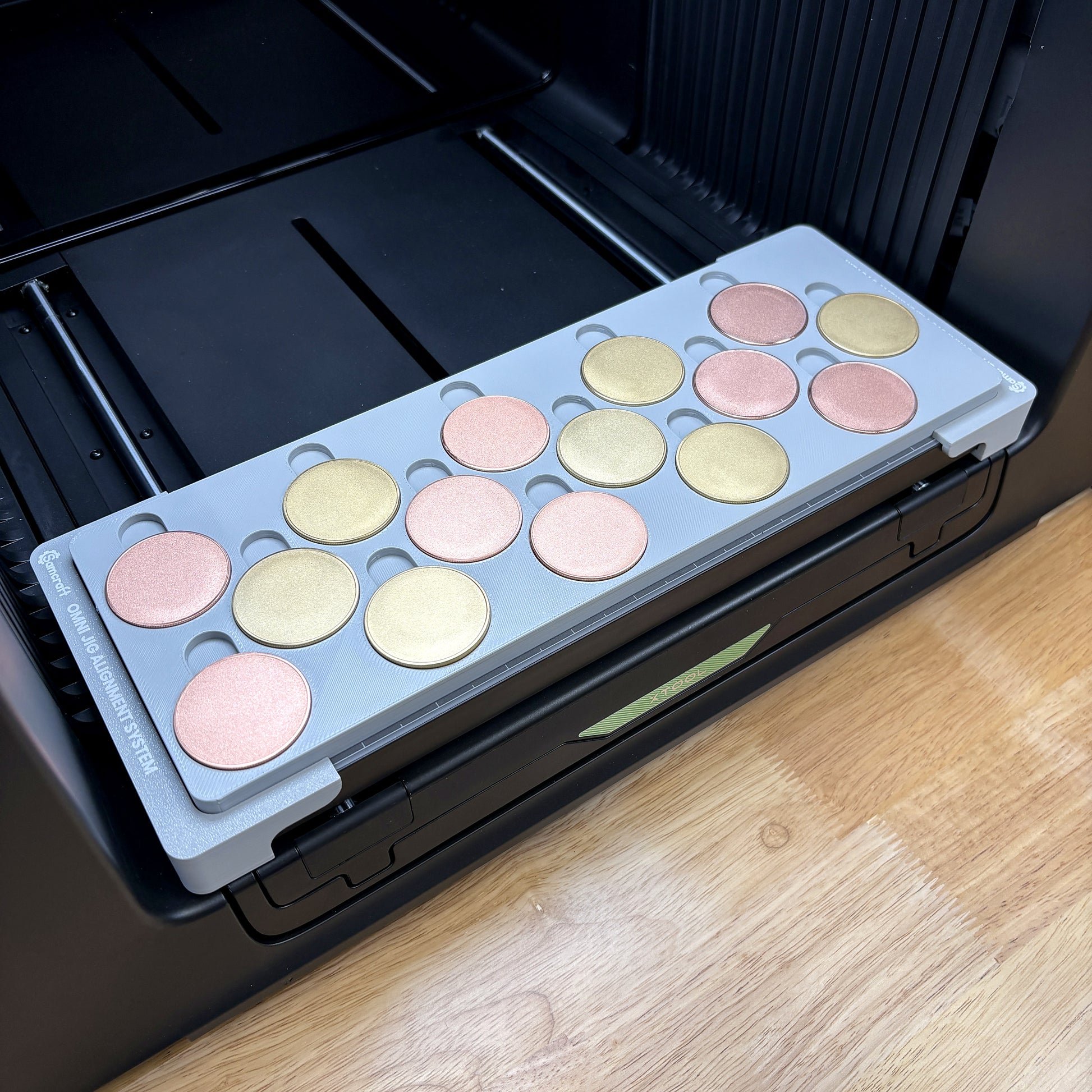 Samcraft 40mm metal coin jig on the bed of an xTool Omni UV printer, holding fifteen rose gold and gold blank challenge coins, seated on the Omni Jig Alignment System.