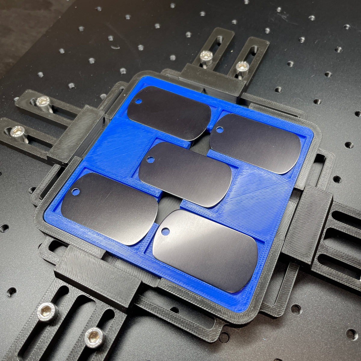 Samcraft blue military dog tag jig on a ComMarker laser bed, holding black and stainless dog tags.