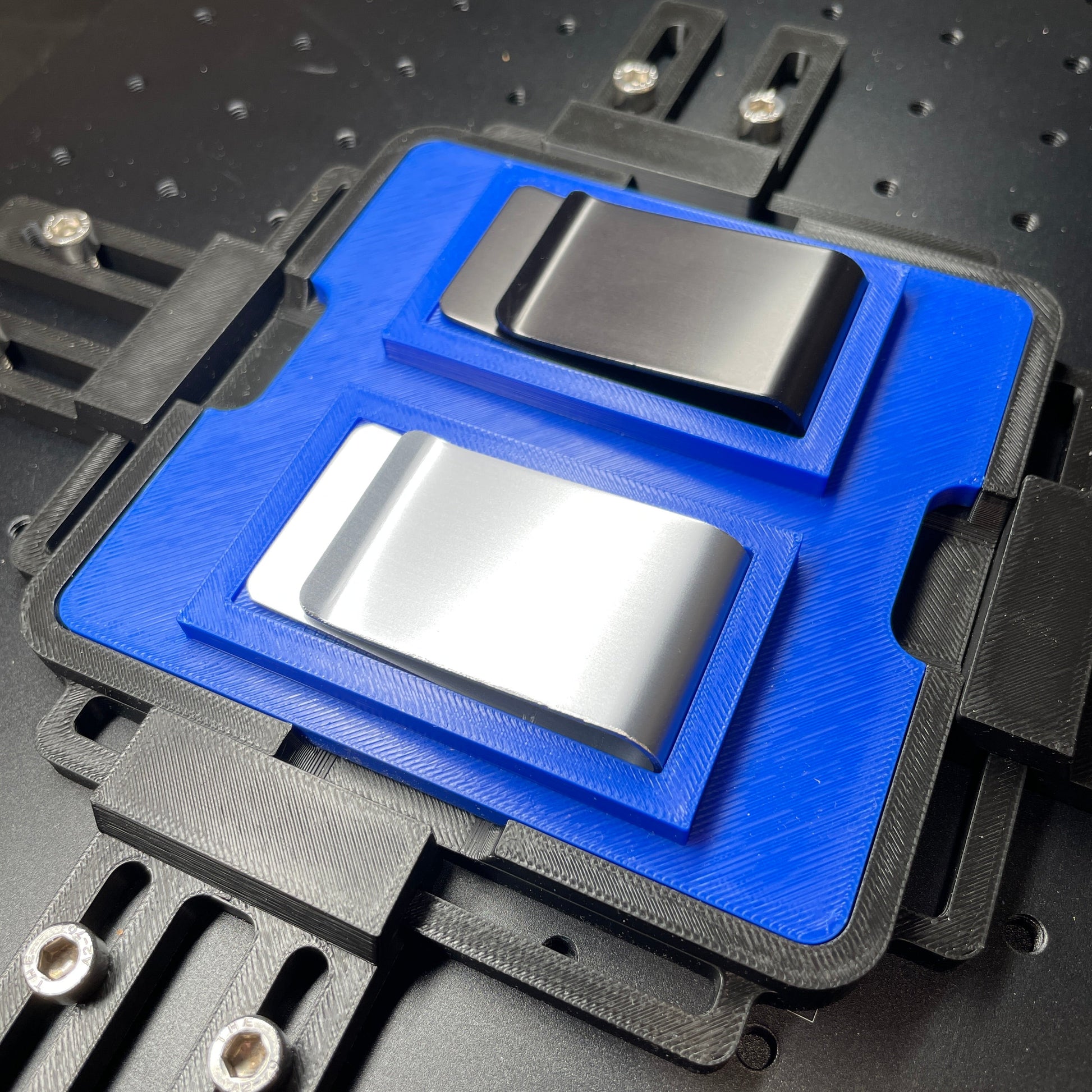 Samcraft blue money clip jig on a ComMarker laser bed, holding a black leatherette money clip and a steel money clip.