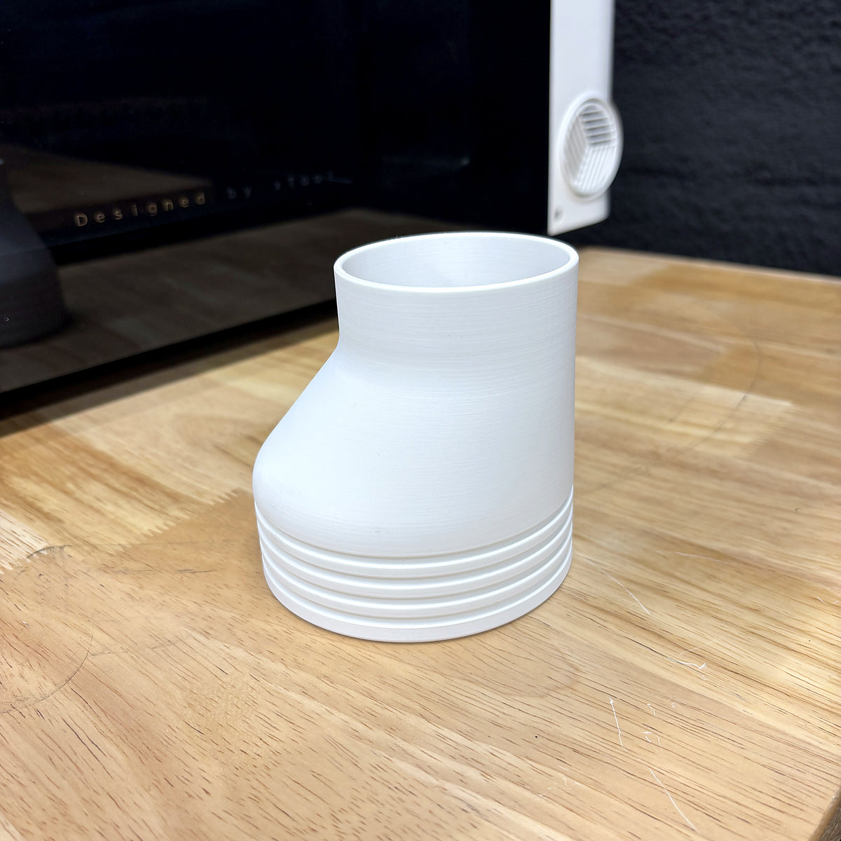 White Samcraft offset 4 inch exhaust hose adapter for the xTool Omni standing on a wood bench in front of the printer, showing the grooved 4 inch end and the smaller port end.