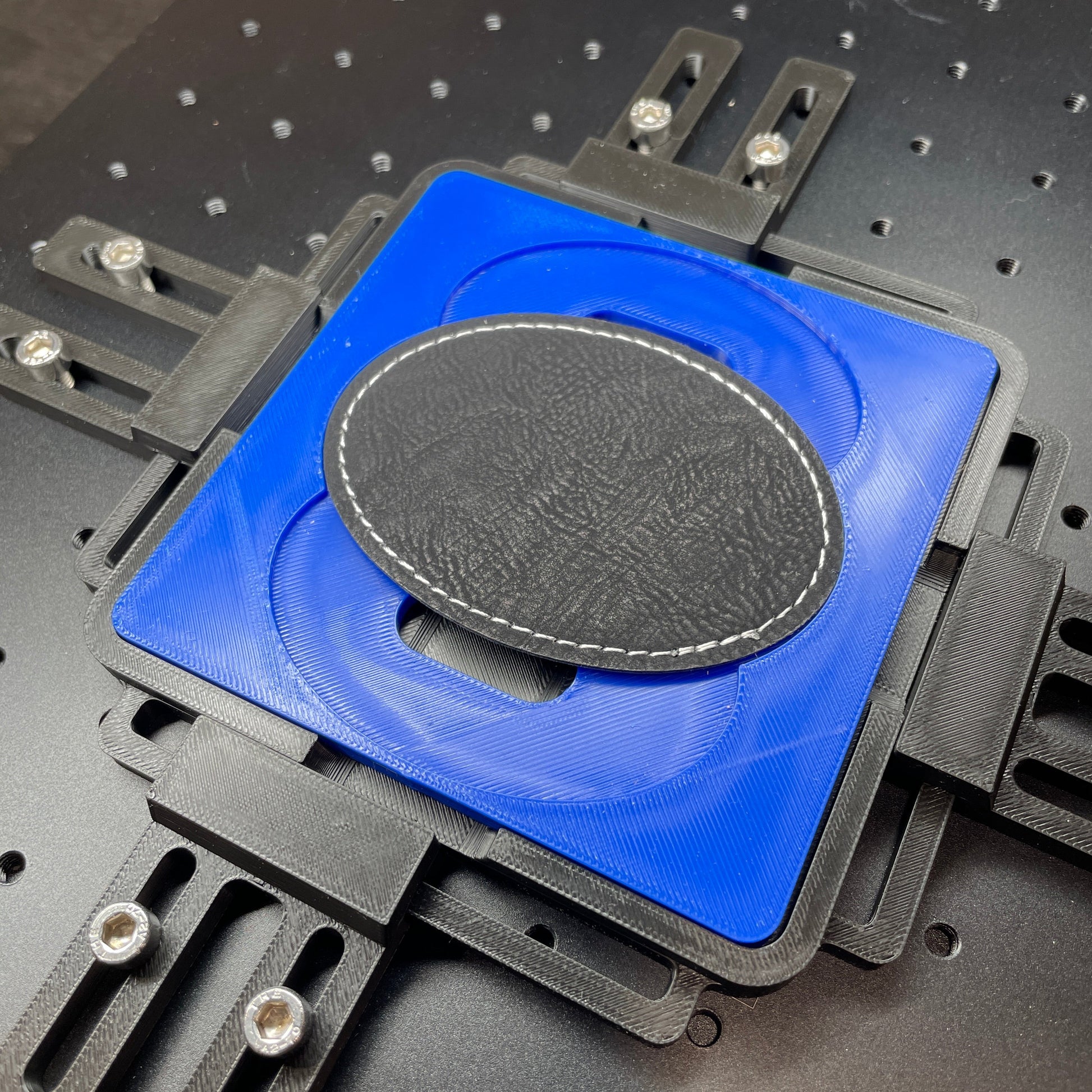 Samcraft blue oval patch jig on a ComMarker laser bed, holding a dark oval leatherette patch.