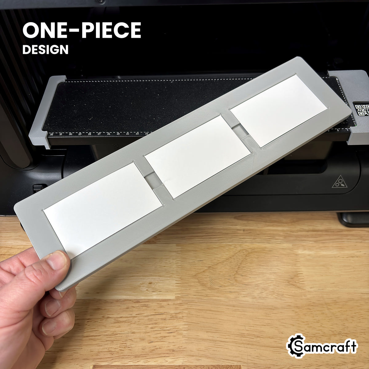 Hand holding the one-piece Samcraft paper business card jig for the eufyMake E1, loaded with three business cards, in front of the printer bed.