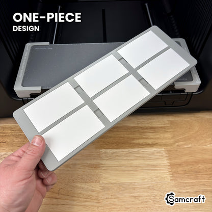 Hand holding the one-piece Samcraft paper business card jig for the xTool Omni, loaded with six business cards, in front of the printer bed.