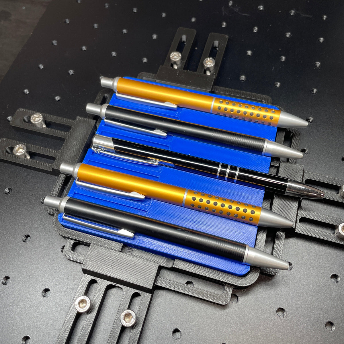 Samcraft blue pen jig on a ComMarker laser bed, holding gold and blue metal pens.