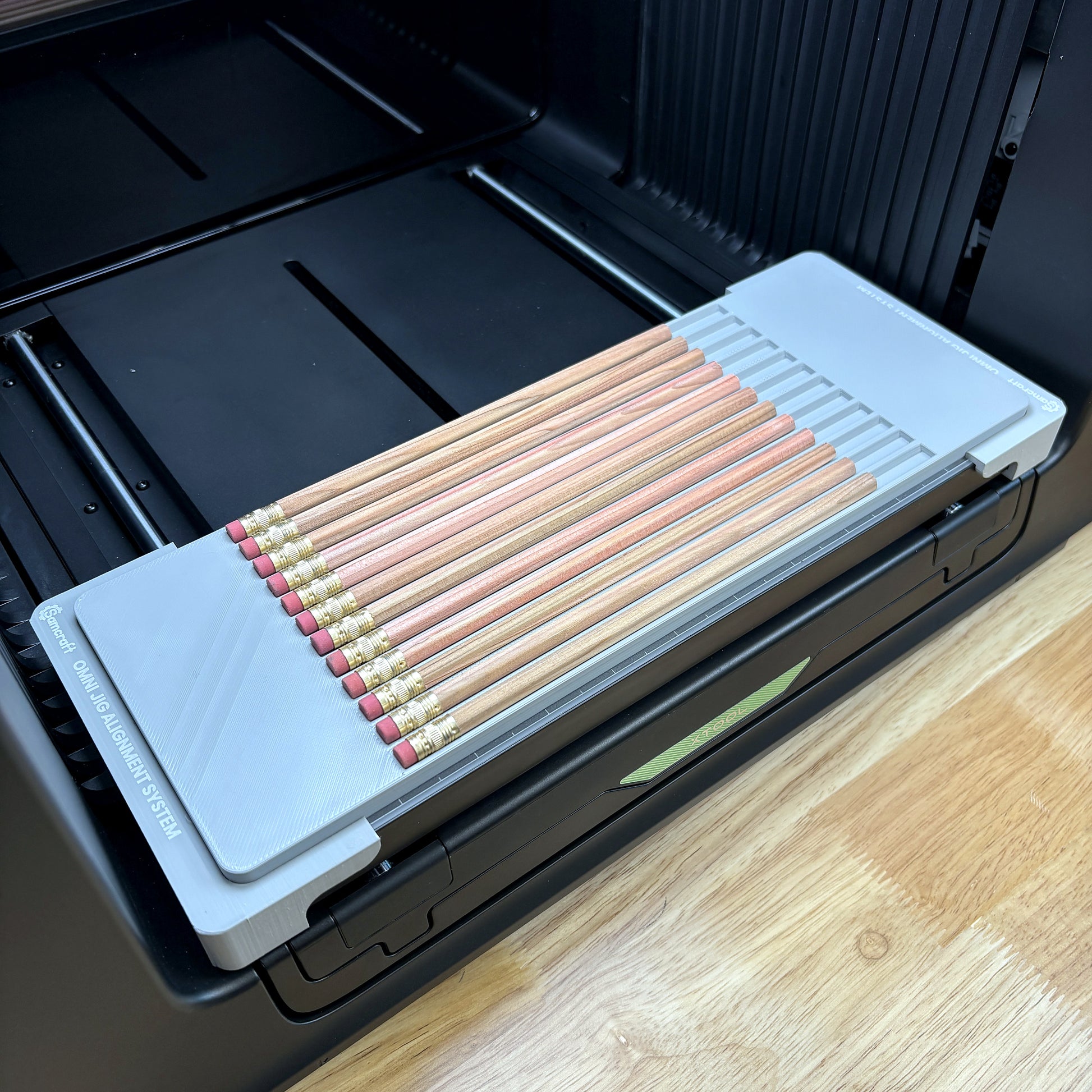 Samcraft hexagon pencil jig on the bed of an xTool Omni UV printer, holding twelve natural wood pencils in parallel channels.