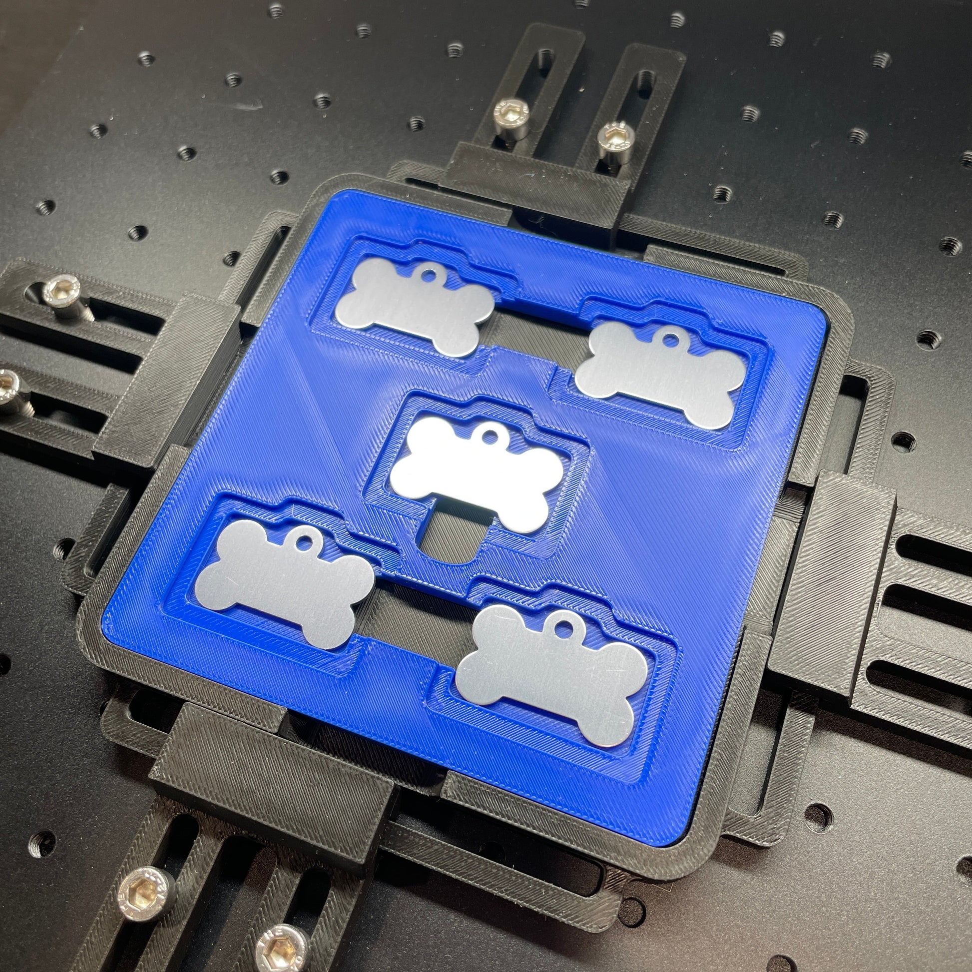 Samcraft blue bone pet tag jig on a ComMarker laser bed, holding stainless bone-shaped pet tags.