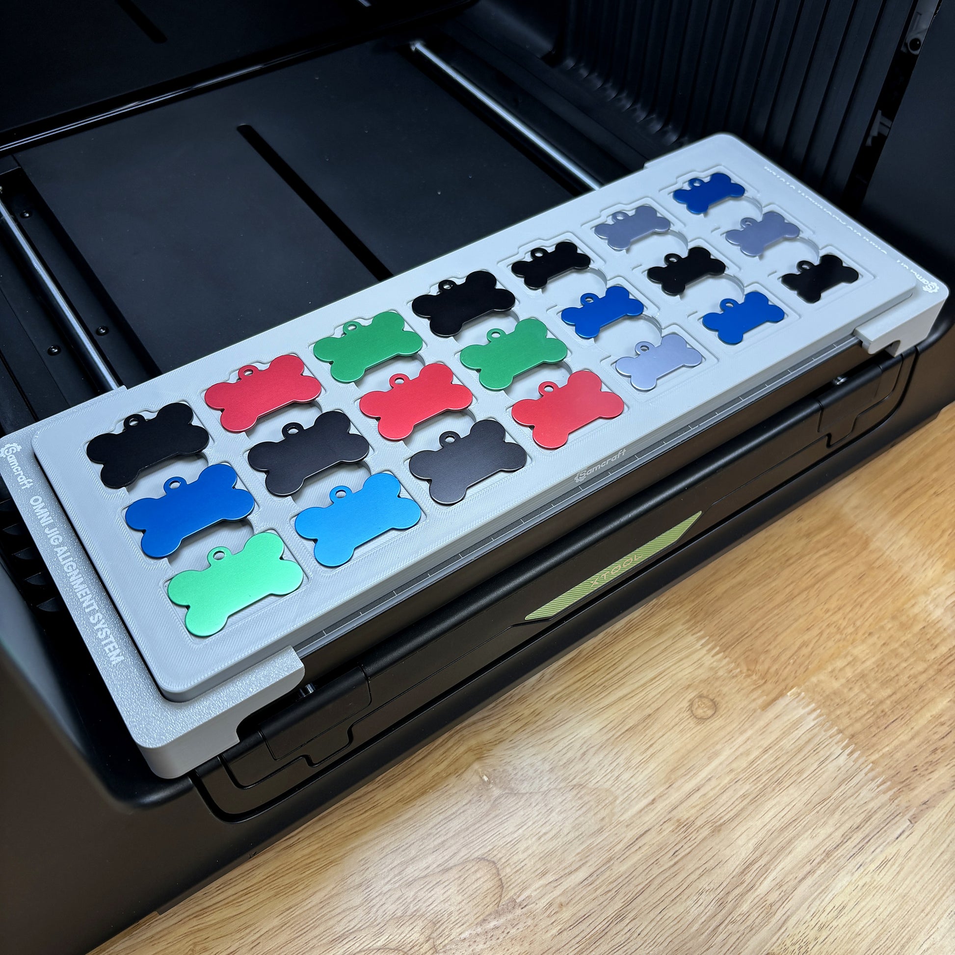 Samcraft bone pet tag jig on the bed of an xTool Omni UV printer, holding 21 anodized aluminum bone tags in red, green, blue, black and silver.