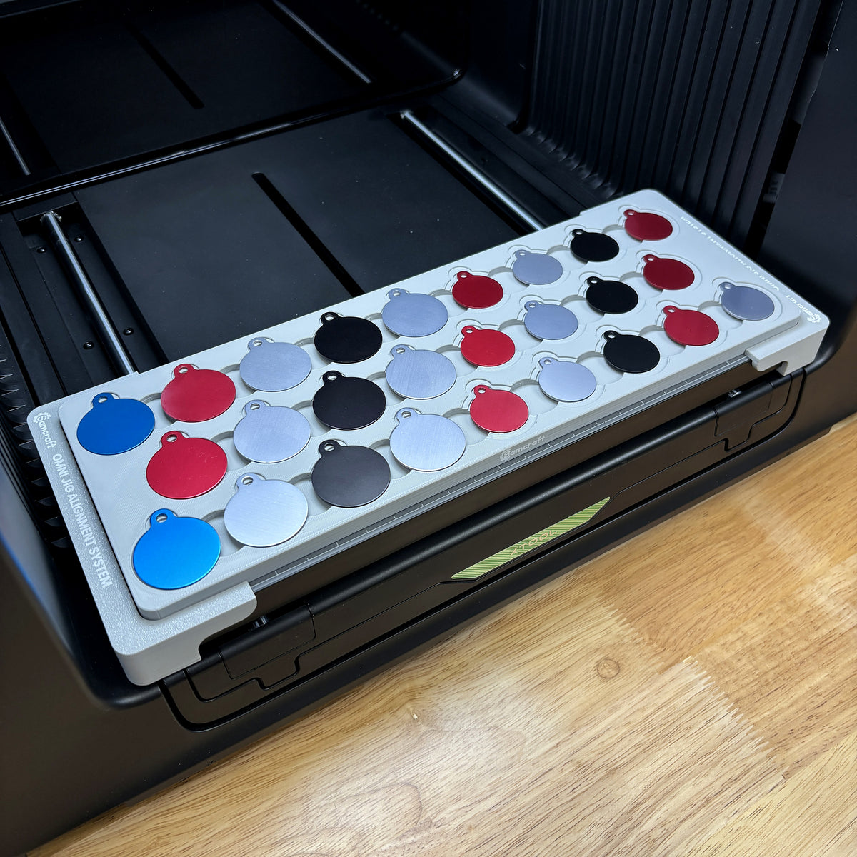 Samcraft round pet tag jig on the bed of an xTool Omni UV printer, holding 26 anodized aluminum round tags in blue, red, silver and black.