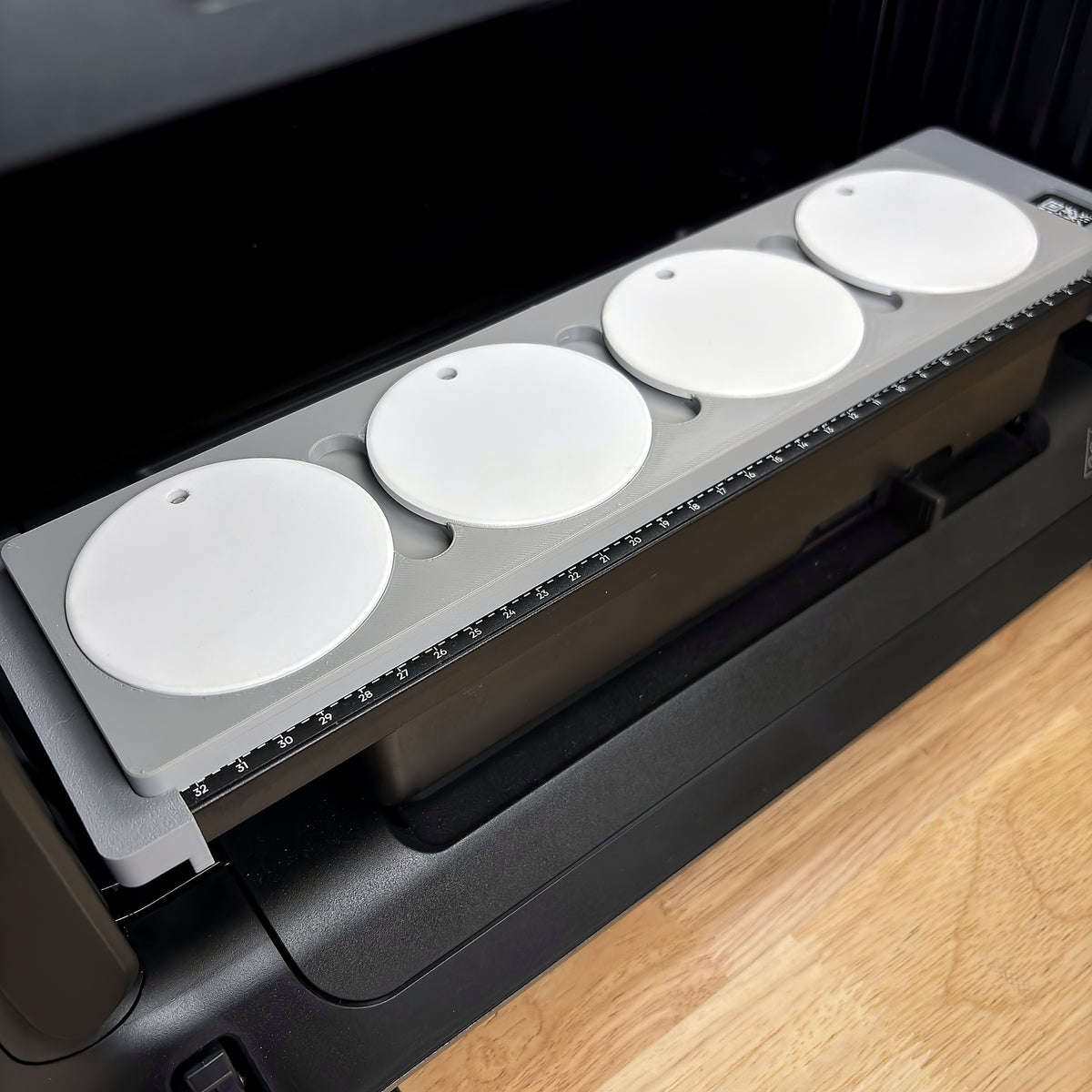 Samcraft round ceramic ornament jig on the bed of a eufyMake E1 UV printer, holding four white round ceramic ornaments in a row beside the bed's built-in ruler.