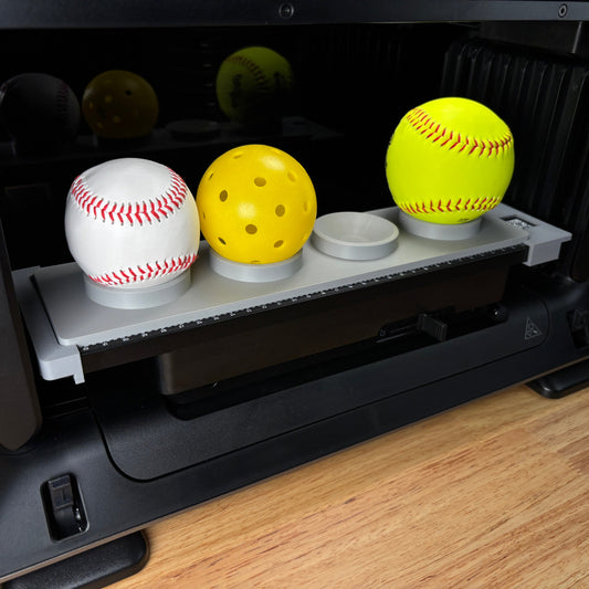 Samcraft sports ball jig on the bed of a eufyMake E1 UV printer, holding a baseball, a yellow pickleball and a yellow softball in cupped recesses.