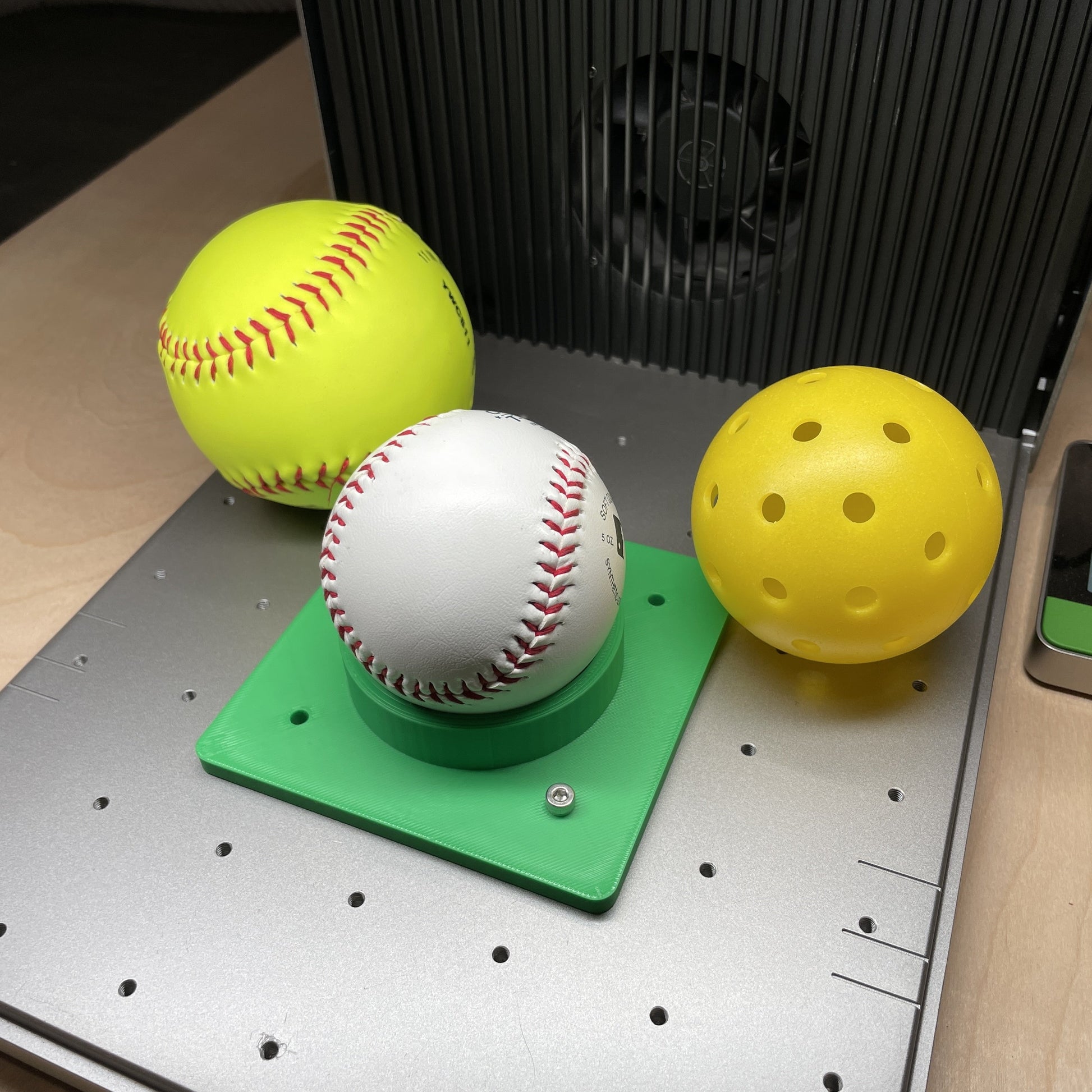 Samcraft green sports ball jig in an xTool F1 Ultra laser bed, cradling a yellow softball, a baseball and a yellow pickleball.