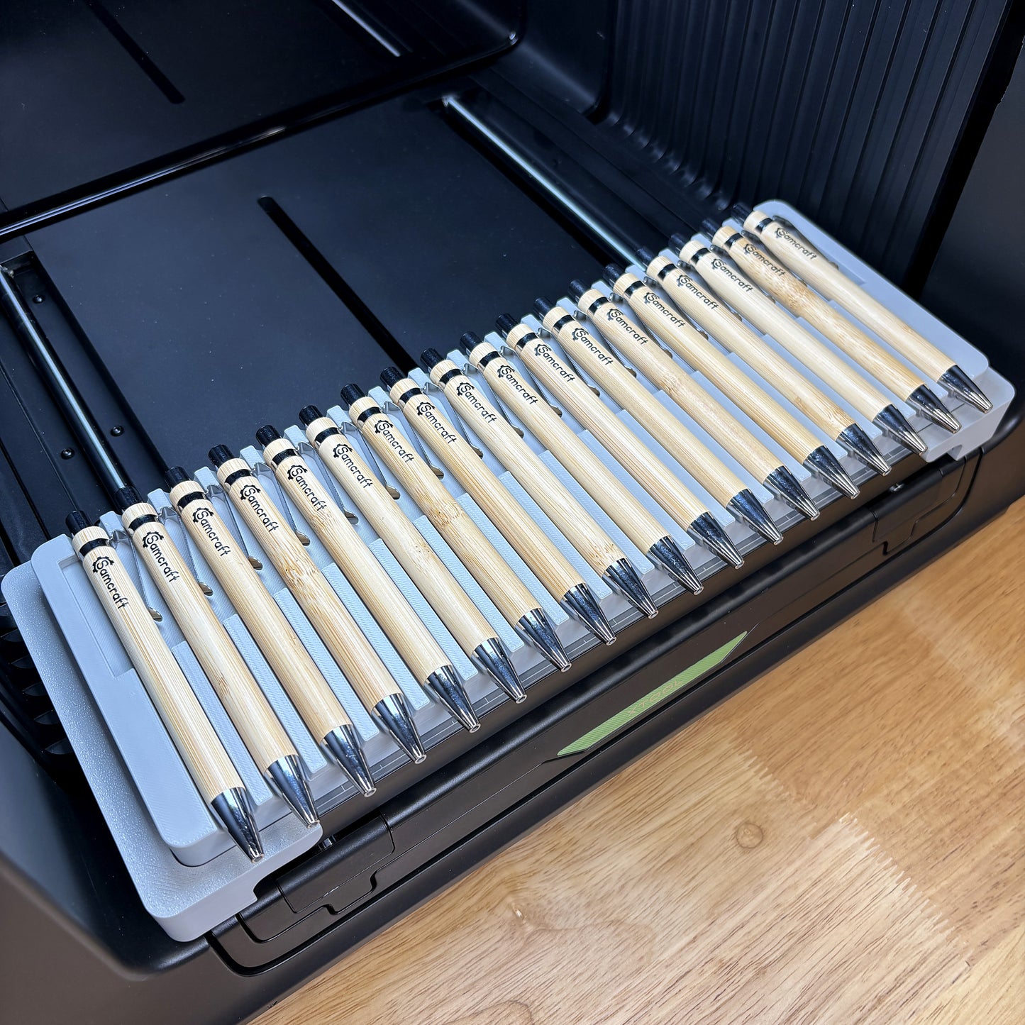 Samcraft universal 18 pen jig on the bed of an xTool Omni UV printer, holding eighteen bamboo pens in curved barrel cradles.