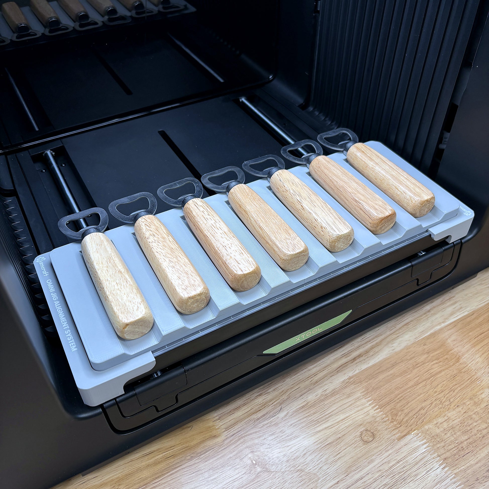 Samcraft wood handle bottle opener jig on the bed of an xTool Omni UV printer, holding seven openers in a single row.